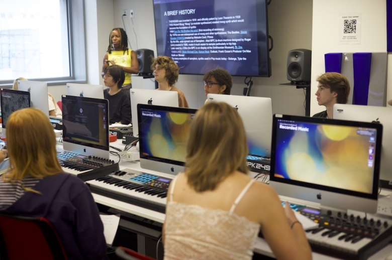 Photo of Recorded Music students in a computer lab sitting in front of Mac desktop computers and MIDI keyboard controllers as they listen to someone speaking out of frame